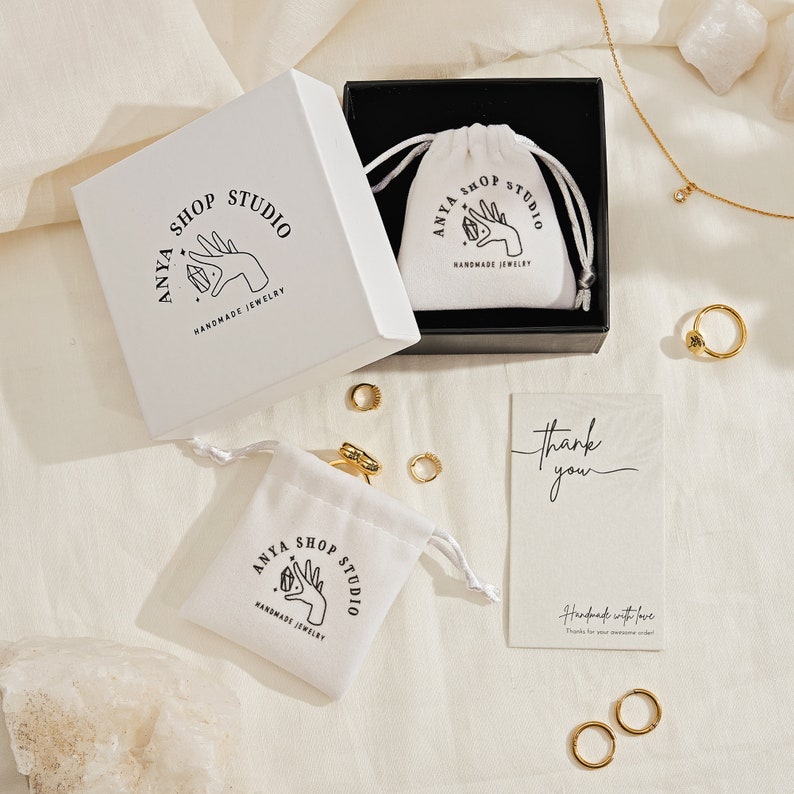 May include: A white jewelry box with a black interior containing a white drawstring pouch, a separate white pouch, several gold rings, a gold necklace with a small pendant, and a thank you card. All items feature the Anya Shop Studio logo.