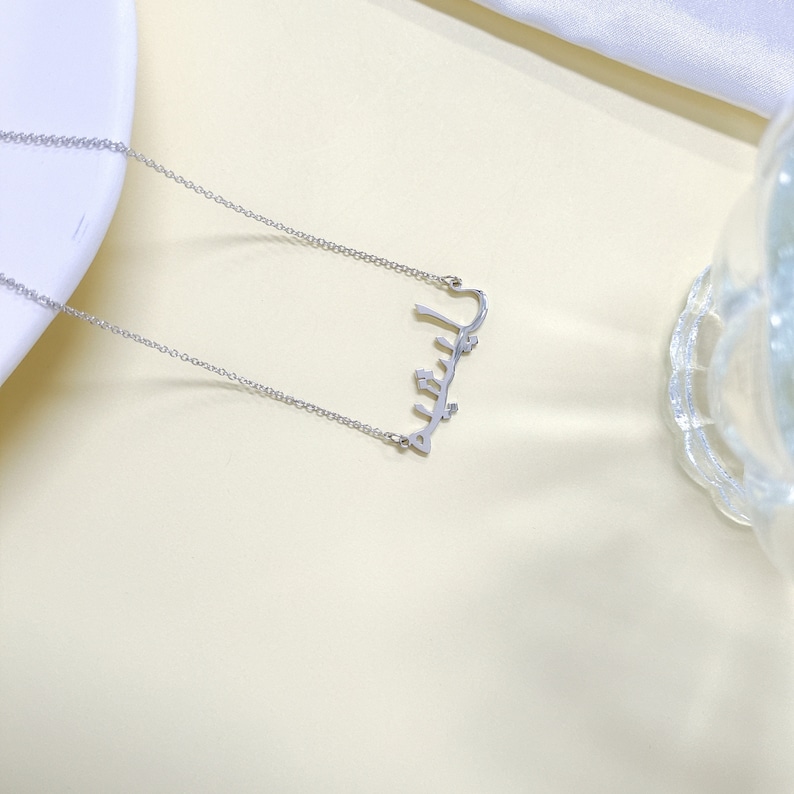 May include: A silver necklace with a delicate chain and a pendant that spells out the word "Jalila" in Arabic script.