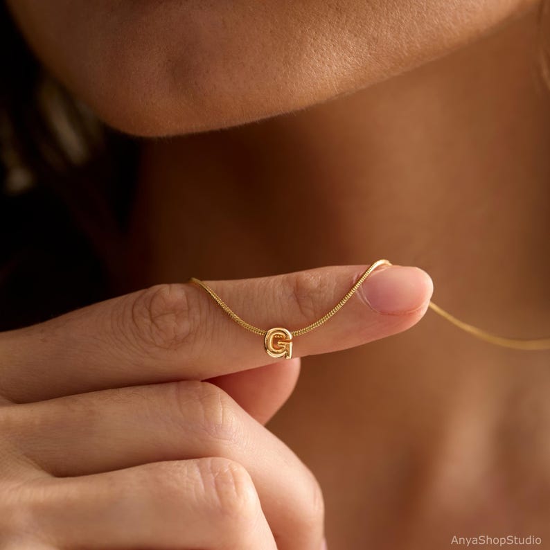 May include: A gold-toned necklace featuring a delicate snake chain and a small, rounded letter G pendant. The pendant is centred on the chain, which rests against the skin. The metal has a polished, reflective finish throughout the piece.
