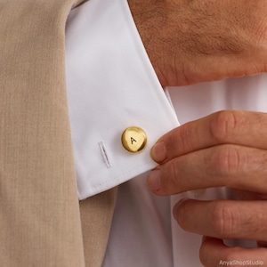 May include: A circular gold-toned cufflink featuring the capital letter A engraved in black on its surface. The cufflink is fastened through the buttonhole of a crisp white shirt cuff, which is partially covered by a tan-colored blazer sleeve.