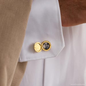 May include: A gold-toned circular locket cufflink attached to a white shirt cuff. The locket is open, revealing a small black and white photograph of two people inside one side, while the other side remains empty and polished.