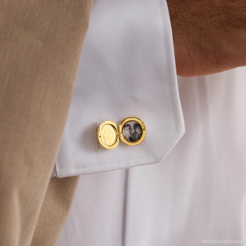 May include: A gold-toned circular locket cufflink attached to a white shirt cuff. The locket is open, revealing a small black and white photograph of two people inside one side, while the other side remains empty and polished.