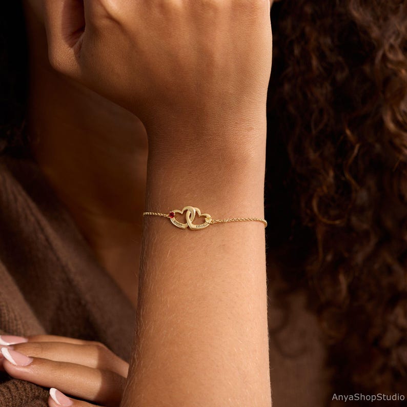 May include: A gold-toned chain bracelet featuring two interlocking heart-shaped charms. One heart is accented with a small red stone, and both hearts are engraved with text. The bracelet is secured with a delicate link chain.