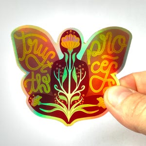 Könnte beinhalten: Ein holografischer Aufkleber mit einem roten, gelben und grünen Blumendesign. Der Aufkleber zeigt eine Person im Lotussitz mit den Worten "Trust the Process" und "Peace" auf beiden Seiten.