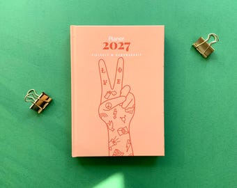 May include: A peach-coloured 2027 planner with a red line art illustration of a hand making a peace sign. The hand features small icons including a peace symbol, lightning bolt, and megaphone. The text reads Planer 2027 and VIELFALT & DANKBARKEIT.