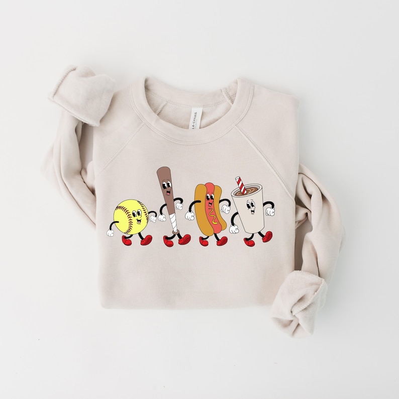May include: A beige sweatshirt with a graphic of a softball, baseball bat, hot dog, and a cup of soda, all with cartoon faces and red shoes.