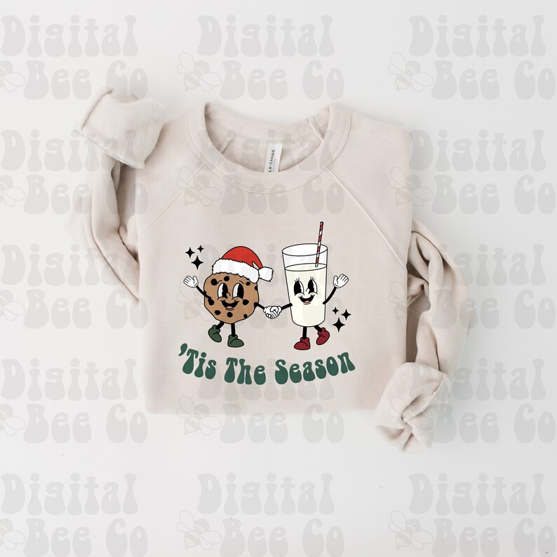 May include: A beige sweatshirt with a Christmas-themed design featuring a smiling cookie and a glass of milk holding hands. The text 'Tis The Season is printed below the design.