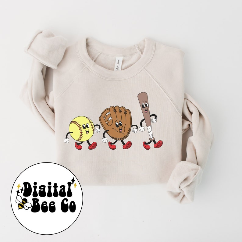 May include: A beige sweatshirt with a graphic of a softball, baseball glove, and baseball bat with cartoon faces and red shoes. The text "Digital Bee Co" is on a yellow and black bee graphic in the lower left corner.