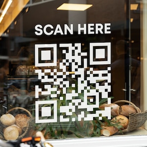 May include: A shop window features a white QR code and the words "SCAN HERE". Inside, baskets of bread and rolls are displayed. The scene suggests a bakery or cafe, inviting customers to scan.