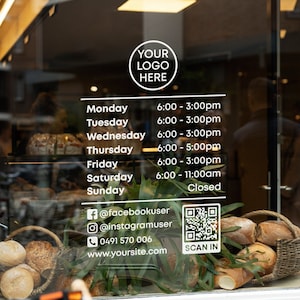 May include: A shop window displaying a sign with daily opening hours. The sign includes the text "YOUR LOGO HERE" and a QR code with the text "SCAN IN". The window also features a basket of bread and a bicycle.