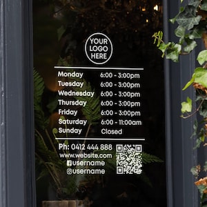 May include: A white sign with black text on a glass door. The sign lists business hours for each day of the week. The sign also includes a phone number, website address, and social media handles. The sign has a QR code in the bottom right corner.