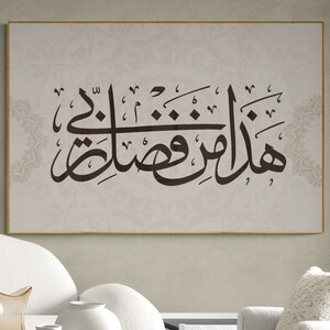 May include: A framed print with Arabic calligraphy in brown ink on a beige background. The print is hanging on a wall above a white couch and a wooden coffee table with white vases.