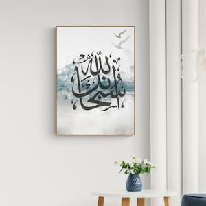 May include: A framed print with a watercolour background in shades of blue and grey. The print features Arabic calligraphy in black, with the text "Allah" in the centre. Two white birds are flying in the top right corner of the print.