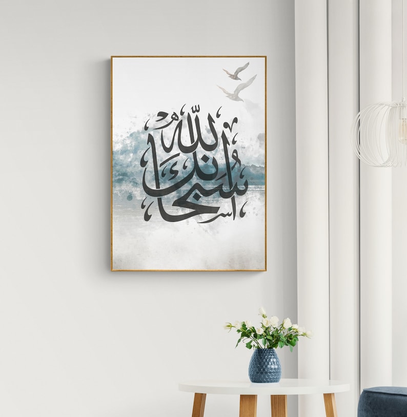 May include: A framed print with a watercolour background in shades of blue and grey. The print features Arabic calligraphy in black, with the text "Allah" in the centre. Two white birds are flying in the top right corner of the print.