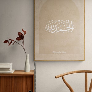 Puede incluir: Una impresi&oacute;n en lienzo beige con un dise&ntilde;o de caligraf&iacute;a &aacute;rabe blanca en forma de arco. El texto dice "Alhamdu Lillah", que se traduce como "Alabado sea Dios".