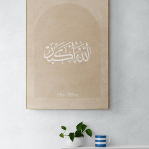 Puede incluir: Una impresi&oacute;n en lienzo beige con un dise&ntilde;o de caligraf&iacute;a &aacute;rabe blanca en forma de arco. El texto dice "Allah Akbar".