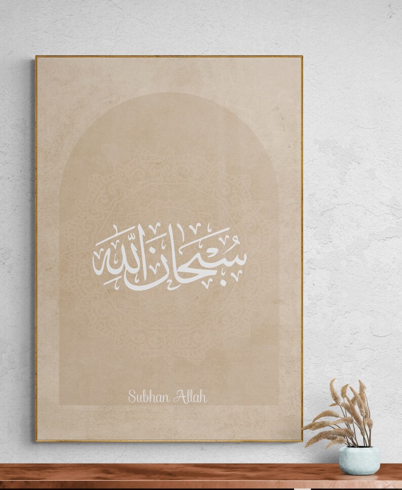 Puede incluir: Una impresi&oacute;n en lienzo beige con marco dorado presenta caligraf&iacute;a &aacute;rabe en blanco, que dice "Subhan Allah" en un arco decorativo. El fondo es un beige sutil con un patr&oacute;n texturizado.