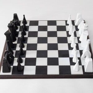 May include: A contemporary chess set featuring black and white pieces on a chequered board. The chess pieces have a geometric design. The board is framed in dark wood. The game is set up and ready to play.