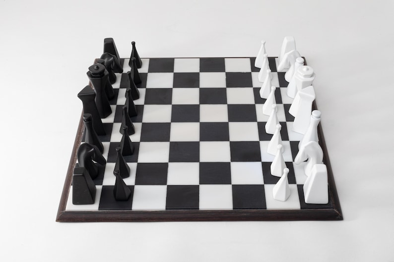 May include: A contemporary chess set featuring black and white pieces on a chequered board. The chess pieces have a geometric design. The board is framed in dark wood. The game is set up and ready to play.