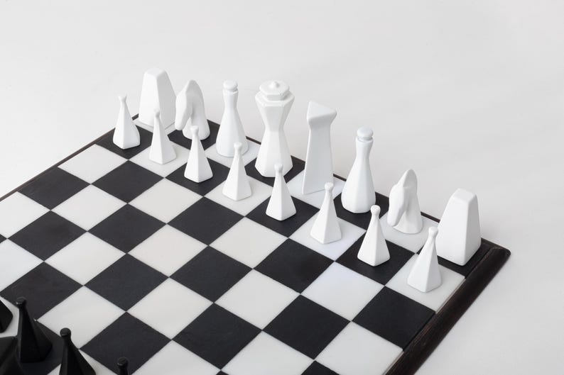 May include: A contemporary chess set featuring white pieces on a black and white chequered board. The chess pieces are minimalist, with geometric shapes and smooth surfaces. The board is framed in dark wood.