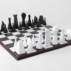 May include: A contemporary chess set featuring black and white pieces on a chequered board. The chess pieces are geometric in design, with black pieces on one side and white on the other. The board has a dark brown border.