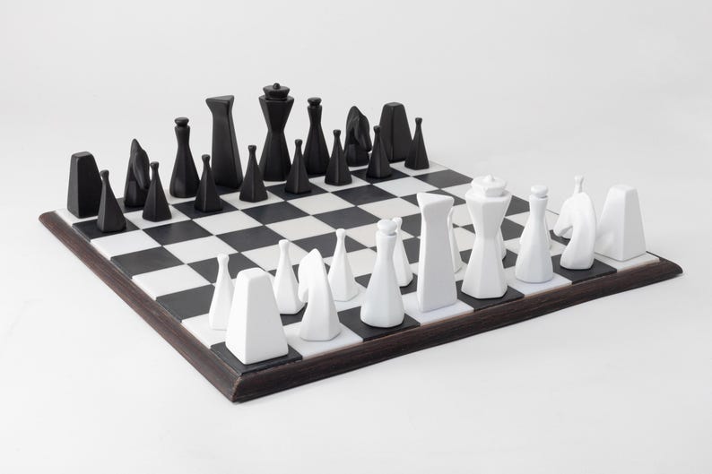 May include: A contemporary chess set featuring black and white pieces on a chequered board. The chess pieces are geometric in design, with black pieces on one side and white on the other. The board has a dark brown border.