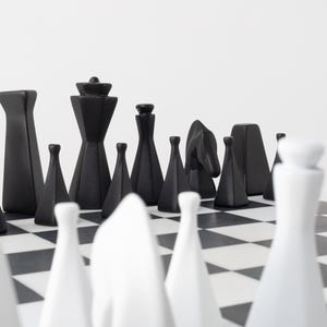 May include: A contemporary chess set featuring black and white pieces on a chequered board. The black pieces are geometric, while the white pieces are more traditional. The pieces are arranged ready to play.