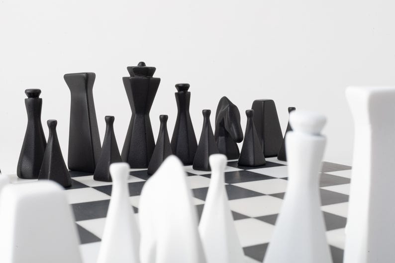 May include: A contemporary chess set featuring black and white pieces on a chequered board. The black pieces are geometric, while the white pieces are more traditional. The pieces are arranged ready to play.