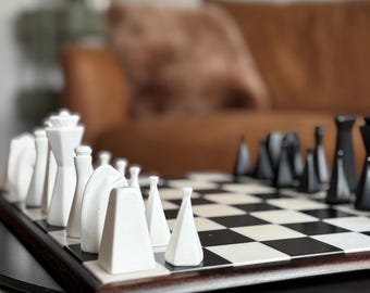 May include: A chess set featuring white and black geometric pieces arranged on a square board with a dark wood frame. The white pieces are positioned in the foreground, showcasing varied angular shapes, while the black pieces are set opposite.