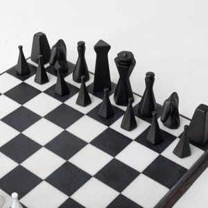 May include: A contemporary chess set featuring black and white pieces on a chequered board. The black pieces are geometric, including cones, cylinders, and a knight. The board is black and white, with a dark wood frame.