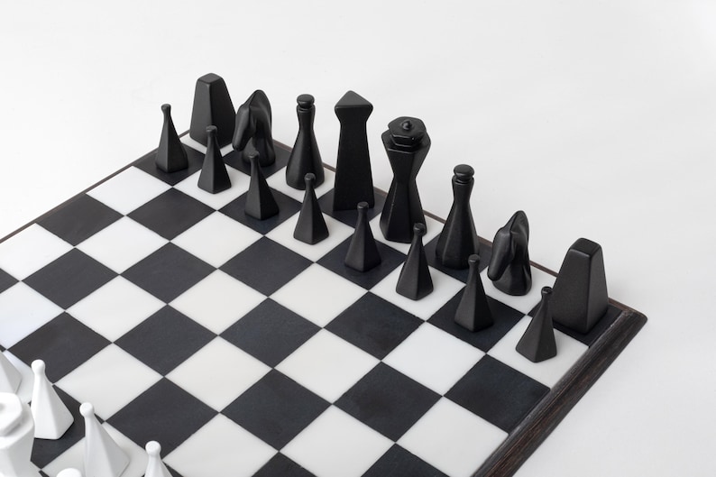 May include: A contemporary chess set featuring black and white pieces on a chequered board. The black pieces are geometric, including cones, cylinders, and a knight. The board is black and white, with a dark wood frame.