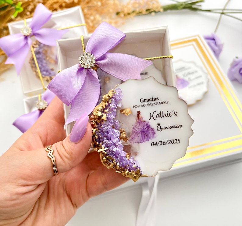 Op de afbeelding: Een ronde, witte, decoratieve gunst met een paars en gouden ontwerp. De gunst toont een paarse jurk met een gouden kroon en de tekst "Gracias por acompa&ntilde;arme Kathie's Quincea&ntilde;era 04/26/2025".