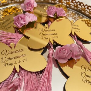 May include: Gold butterfly-shaped mirror favors with pink tassels and flowers. The mirrors are personalized with the text "Vanessa Quincea&ntilde;era May 2, 2022".