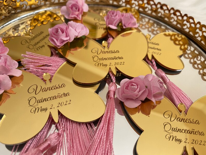 May include: Gold butterfly-shaped mirror favors with pink tassels and flowers. The mirrors are personalized with the text "Vanessa Quincea&ntilde;era May 2, 2022".