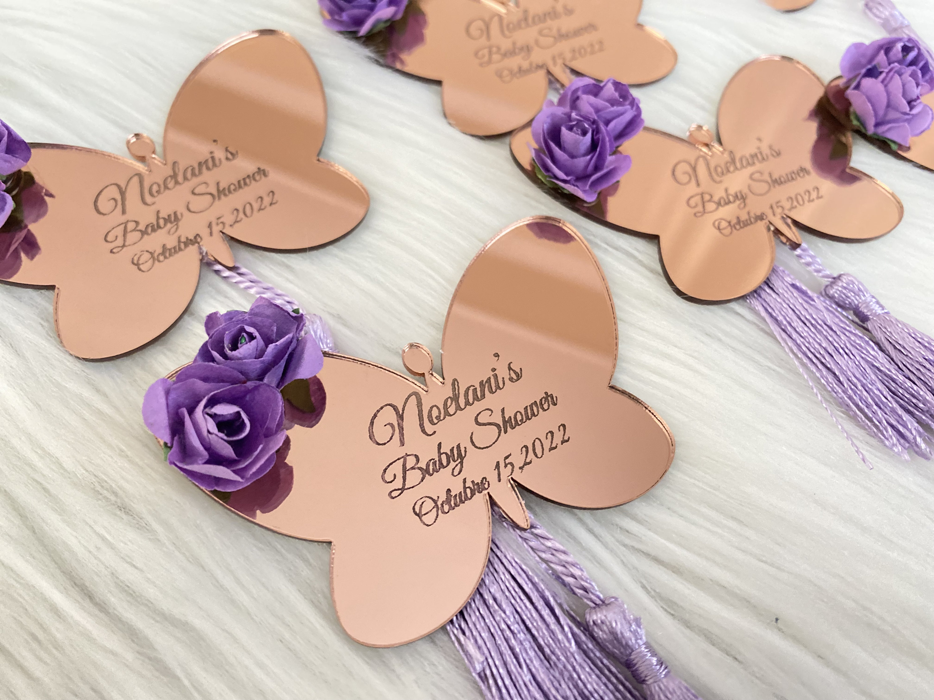 May include: Rose gold butterfly shaped mirror favors with purple flowers and tassels. The favors are personalized with the text "Noelani's Baby Shower October 15, 2022".