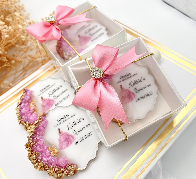 Op de afbeelding: Roze en gouden cadeaudoosjes met een witte achtergrond en een gedrukt beeld van een meisje in een roze jurk. De doosjes hebben een roze lint en een doorzichtig deksel. De tekst op de doosjes luidt "Gracias por acompa&ntilde;arme Kathie's Quincea&ntilde;era 04/26/2025".