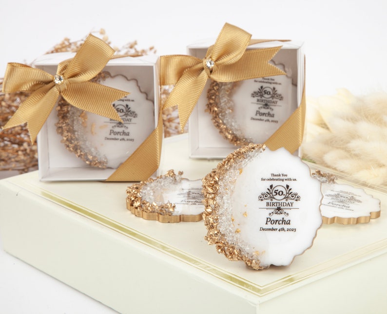 Puede incluir: Cajas de regalo doradas y blancas con un lazo dorado y un posavasos redondo blanco con purpurina dorada y el texto "50. Cumplea&ntilde;os Porcha 4 de diciembre de 2023"