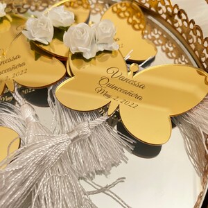 May include: Gold butterfly-shaped mirror favors with white tassels and white flowers. The mirror has the text "Vanessa Quincea&ntilde;era May 2, 2022" engraved on it.