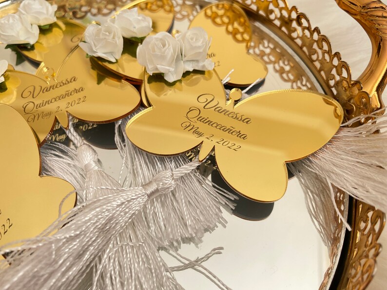May include: Gold butterfly-shaped mirror favors with white tassels and white flowers. The mirror has the text "Vanessa Quincea&ntilde;era May 2, 2022" engraved on it.