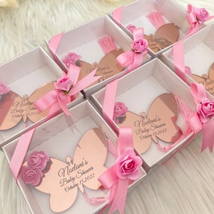 May include: Pink and white gift boxes with rose-gold butterfly shaped favors. Each favor has a pink tassel and the text "Noelani's Baby Shower October 15, 2022".