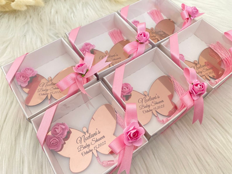 May include: Pink and white gift boxes with rose-gold butterfly shaped favors. Each favor has a pink tassel and the text "Noelani's Baby Shower October 15, 2022".