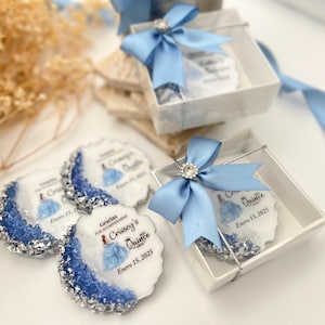 Op de afbeelding: Blauwe en zilveren cadeauboxen met een witte achtergrond. De dozen bevatten ronde, witte onderzetters met een blauw en zilver ontwerp en de tekst "Gracias por acompa&ntilde;arme Crissey's Quince Enero 15, 2025".