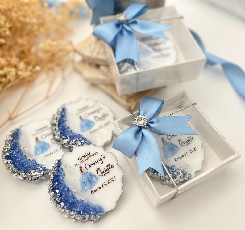 Op de afbeelding: Blauwe en zilveren cadeauboxen met een witte achtergrond. De dozen bevatten ronde, witte onderzetters met een blauw en zilver ontwerp en de tekst "Gracias por acompa&ntilde;arme Crissey's Quince Enero 15, 2025".