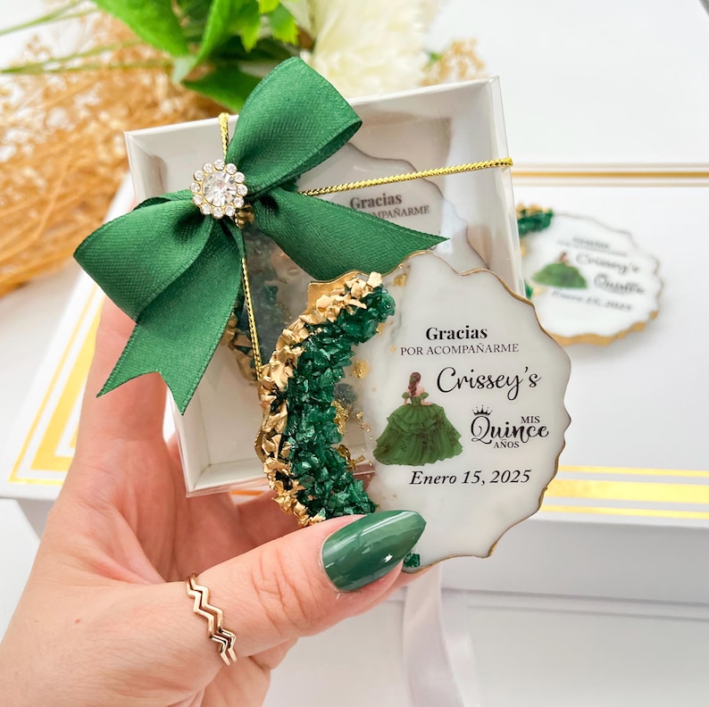 Op de afbeelding: Een witte en gouden ronde gunst met een groen en gouden ontwerp. De gunst toont een meisje in een groene jurk en de tekst "Gracias por acompa&ntilde;arme Crissey's Quince Mis A&ntilde;os Enero 15, 2025".