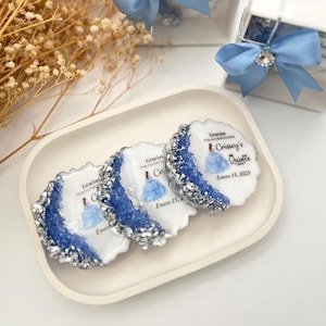 Op de afbeelding: Drie ronde, witte gunsten met blauwe en zilveren glitteraccenten. Elke gunst heeft een gedrukt beeld van een meisje in een blauwe jurk en de tekst "Gracias por acompa&ntilde;ar Crissey's Quince Enero 15, 2025".