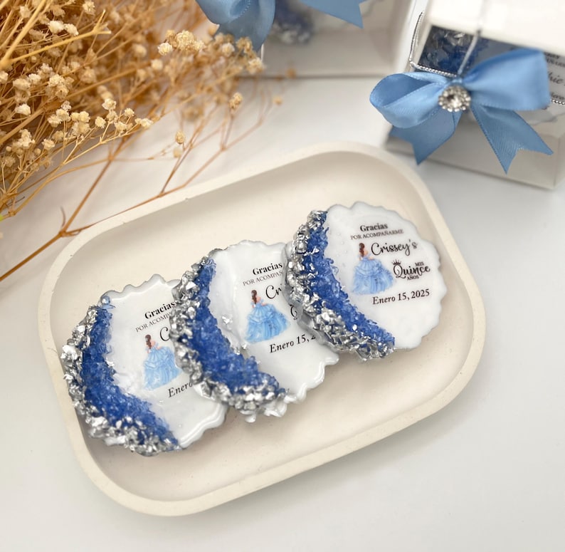 Op de afbeelding: Drie ronde, witte gunsten met blauwe en zilveren glitteraccenten. Elke gunst heeft een gedrukt beeld van een meisje in een blauwe jurk en de tekst "Gracias por acompa&ntilde;ar Crissey's Quince Enero 15, 2025".