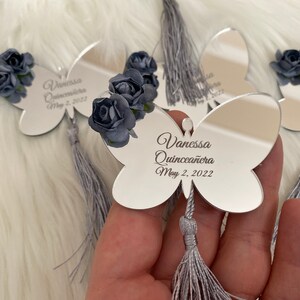 May include: Silver butterfly-shaped mirror with a gray tassel. The mirror has the text "Vanessa Quincea&ntilde;era May 2, 2022" engraved on it. The mirror is decorated with three blue fabric flowers.