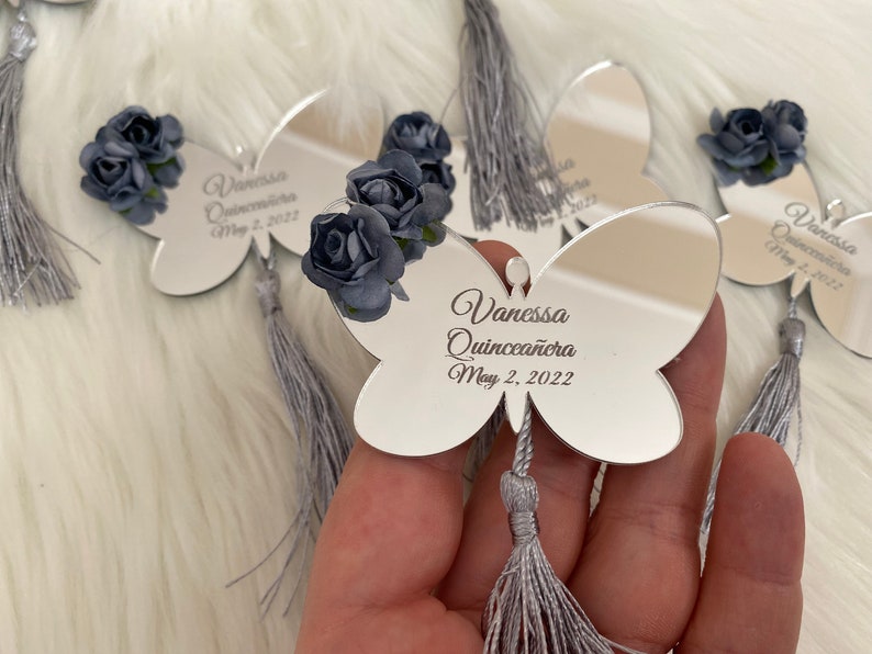 May include: Silver butterfly-shaped mirror with a gray tassel. The mirror has the text "Vanessa Quincea&ntilde;era May 2, 2022" engraved on it. The mirror is decorated with three blue fabric flowers.
