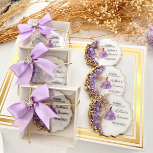 Op de afbeelding: Paarse en gouden cadeaudoosjes met een witte achtergrond. Elk doosje bevat een ronde, witte koek met een paars en gouden ontwerp en de tekst "Gracias por acompa&ntilde;arme Kathie's Quincea&ntilde;era 04/26/2025".