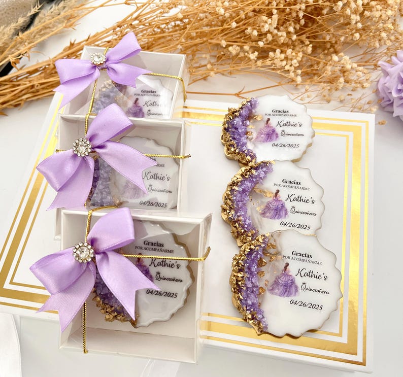 Op de afbeelding: Paarse en gouden cadeaudoosjes met een witte achtergrond. Elk doosje bevat een ronde, witte koek met een paars en gouden ontwerp en de tekst "Gracias por acompa&ntilde;arme Kathie's Quincea&ntilde;era 04/26/2025".